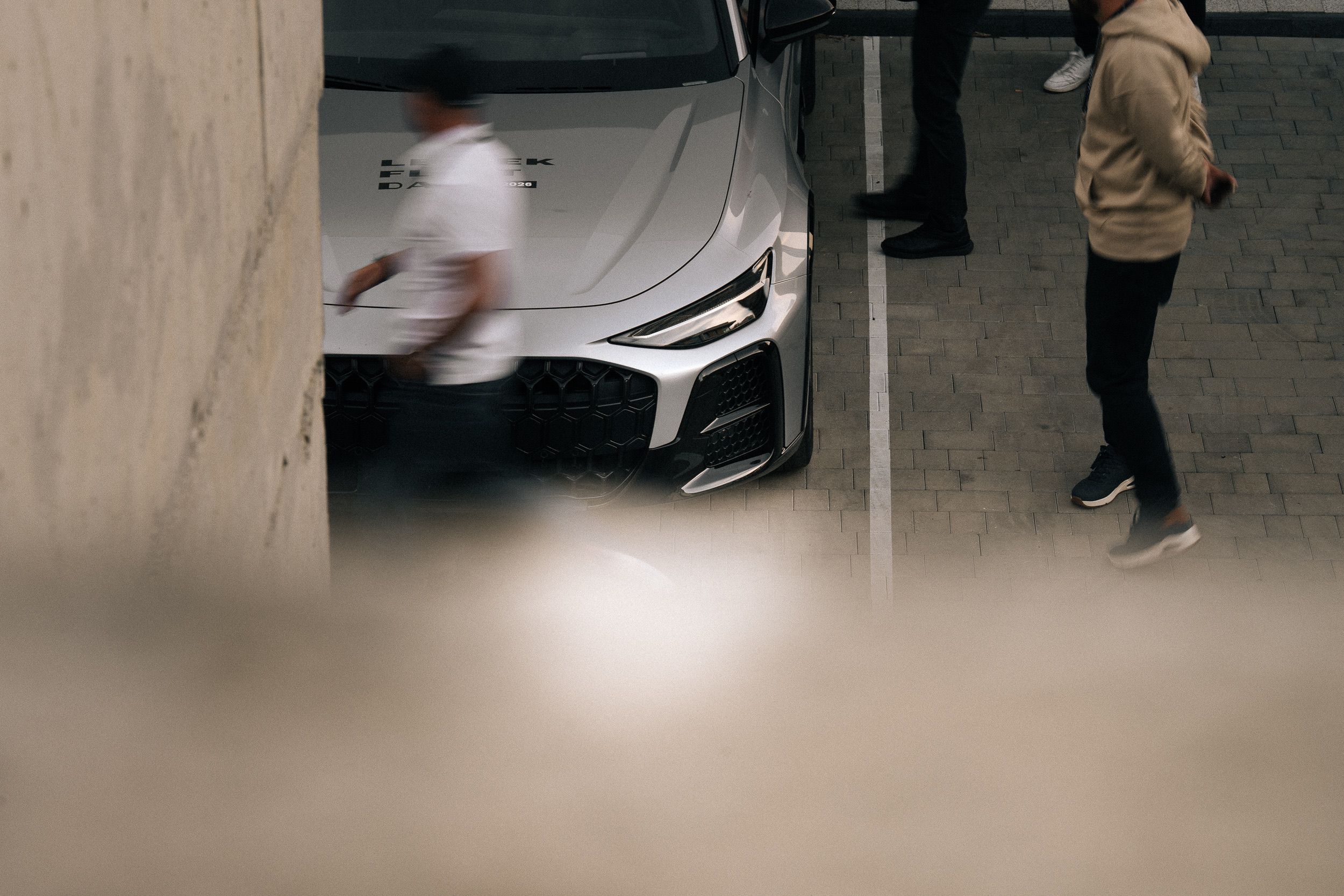 Kadr zza ściany: goście skupieni wokół srebrnego Audi na parkingu