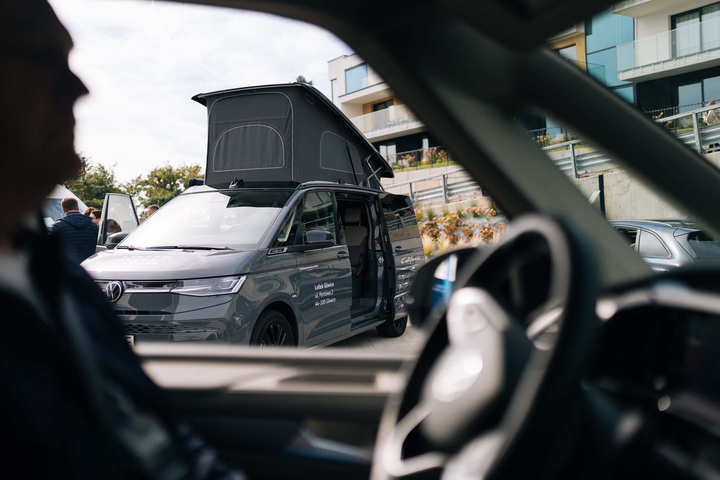 Z kabiny: sylwetka kierowcy i kierownica, za szybą VW California z podniesionym dachem i otwartymi drzwiami przy hotelu